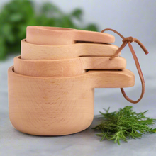 Load image into Gallery viewer, A set of natural wood measuring cups in varying sizes, designed to nest, with a beige wood grain finish and a small strap for hanging storage.

